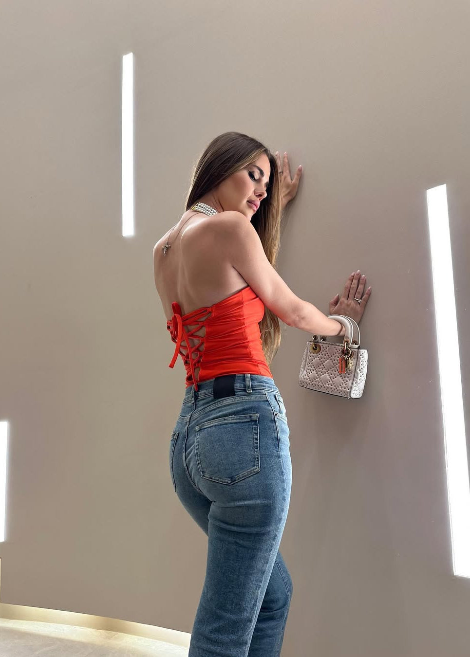 Woman in red top and blue jeans holding a handbag against a neutral wall.