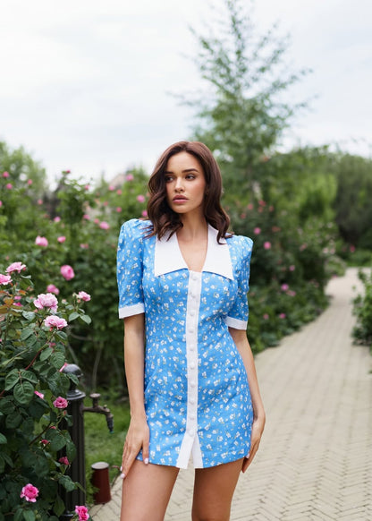 Woman in a blue dress with white polka dots standing in a garden with pink flowers.