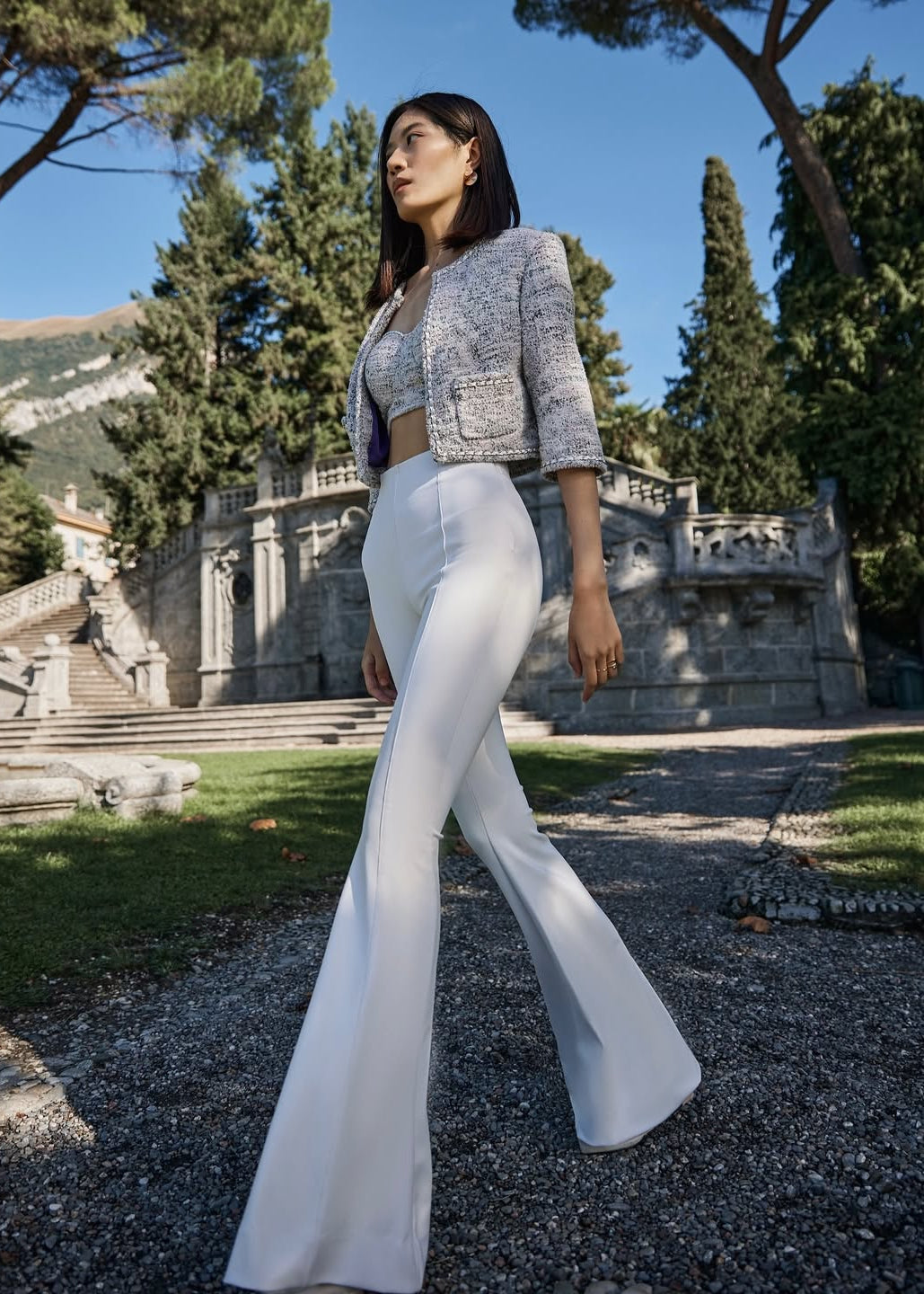 Woman in a stylish outfit standing in a scenic outdoor setting with trees and architecture.