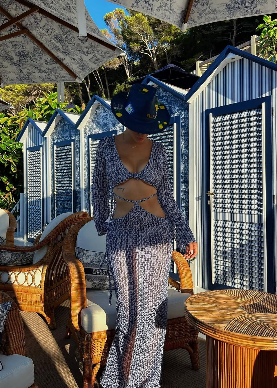 Person wearing a blue patterned dress and hat in an outdoor setting with wicker furniture and beach huts.
