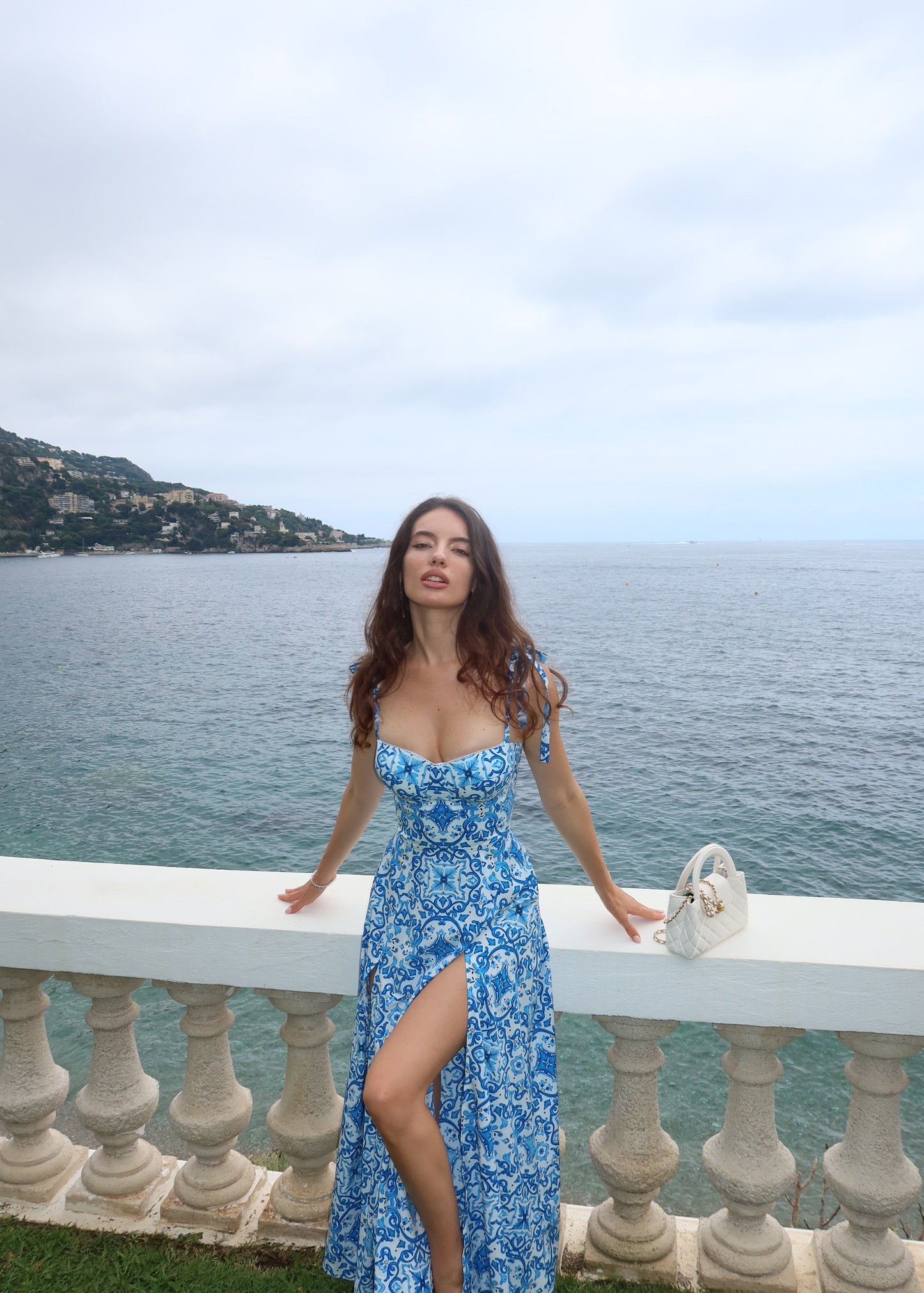 Woman in a blue dress standing on a balcony overlooking a scenic view of water and mountains.