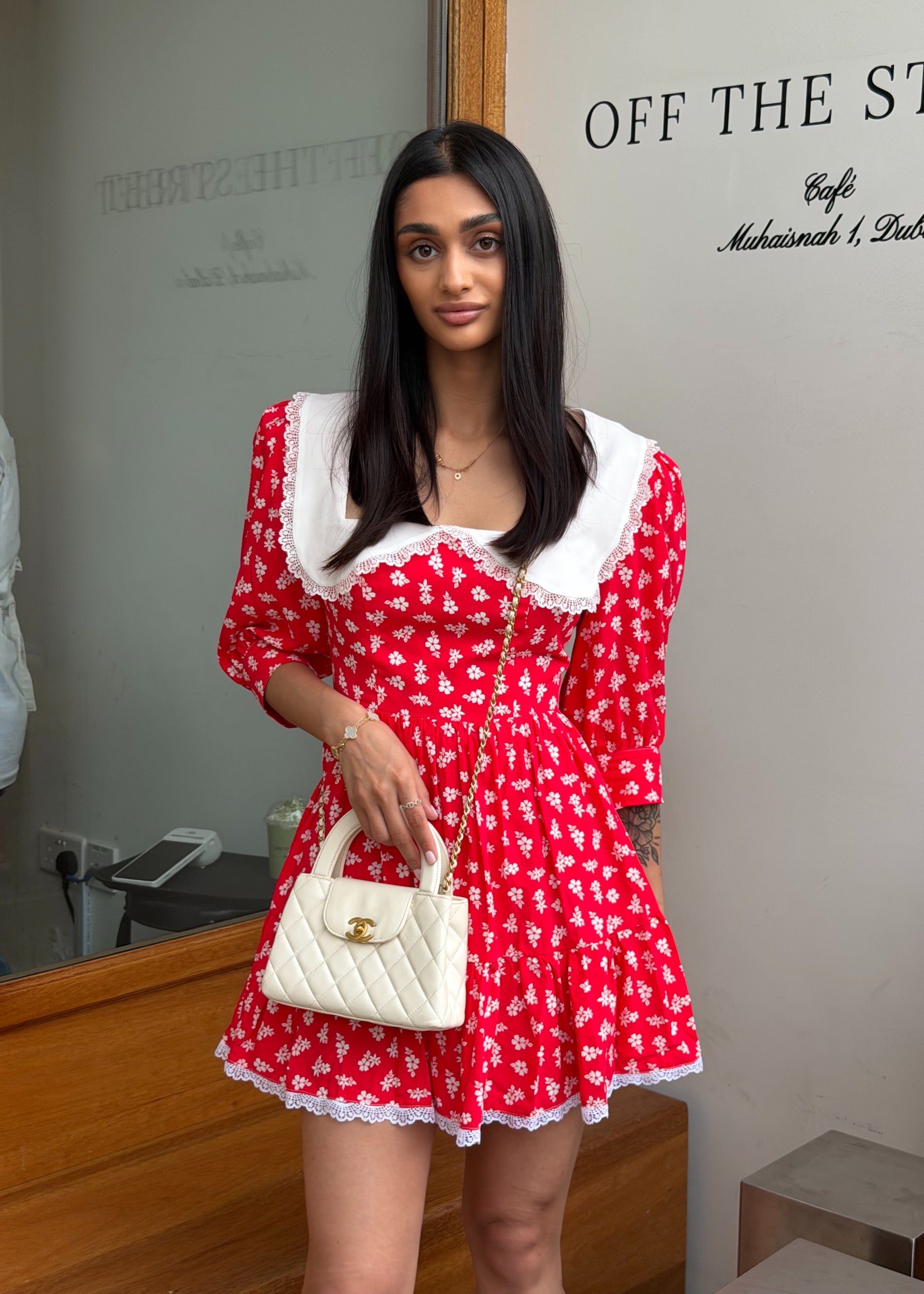 Woman in a red dress with white floral pattern holding a white handbag, standing in front of a mirror.