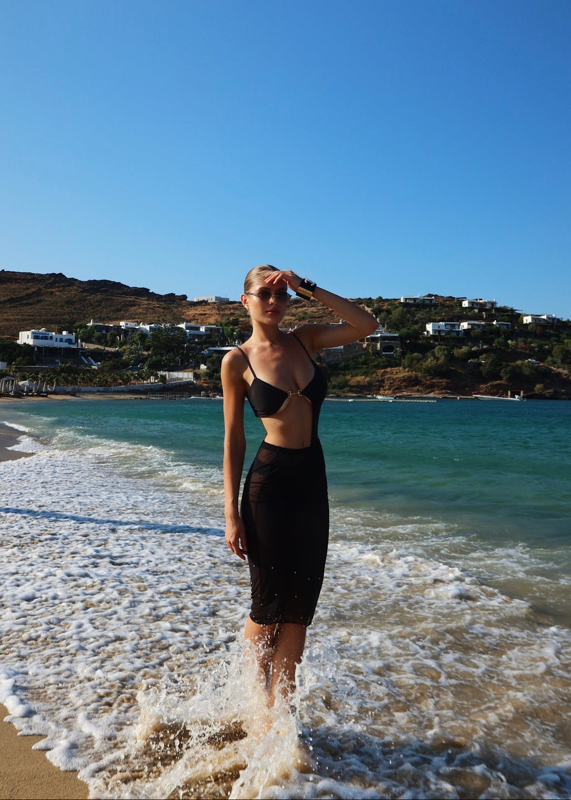 Woman in a black dress standing on a beach with clear blue sky and ocean.
