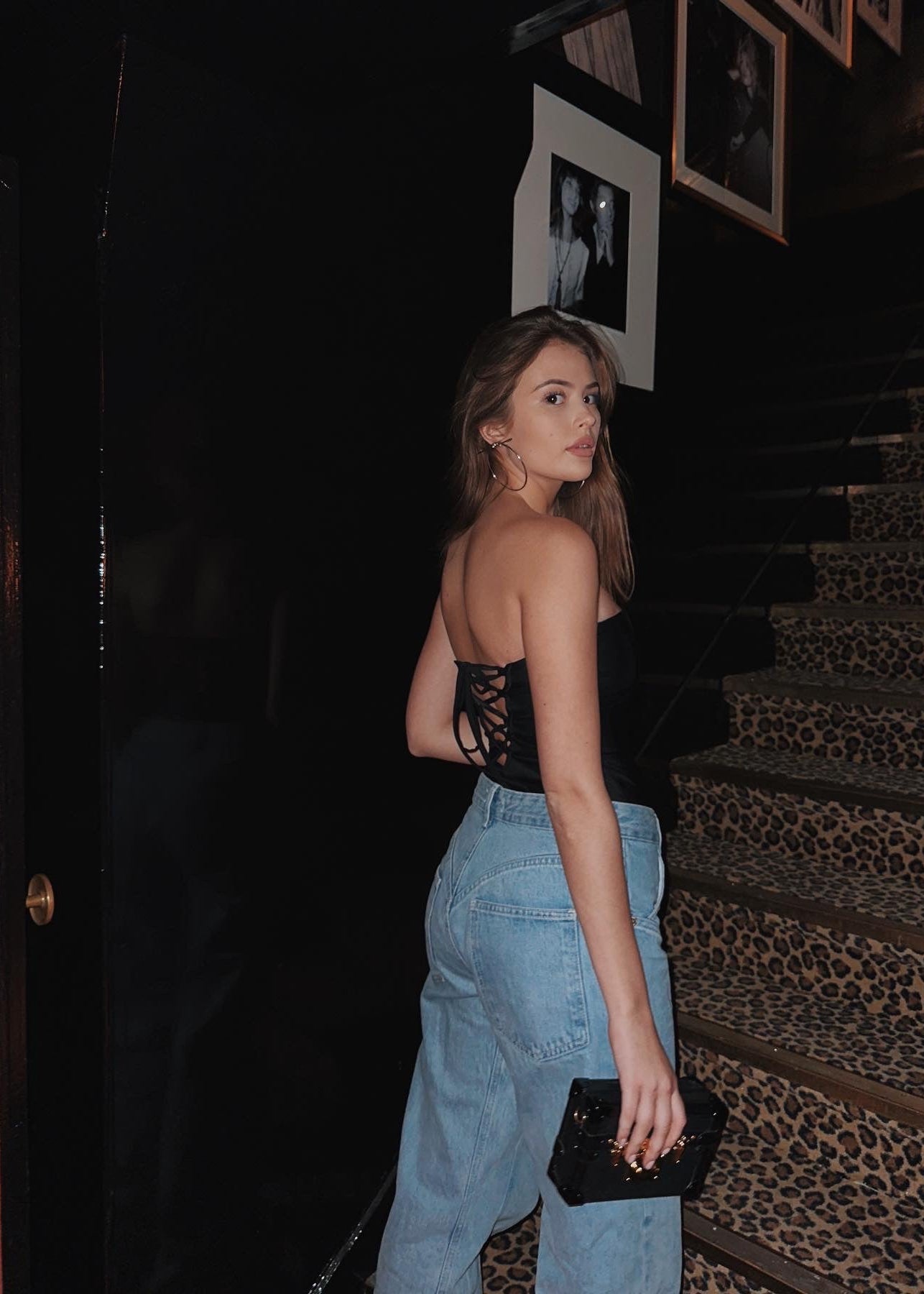 Woman in black top and blue jeans standing on a staircase with a black clutch.