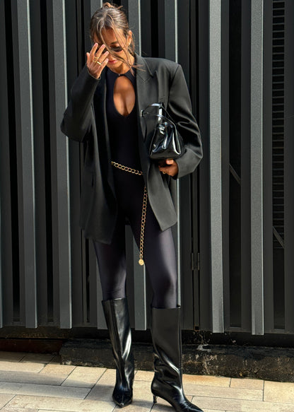 Person wearing a black outfit with a long coat and high boots, holding a black bag against a dark background.