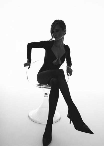 Woman in a black bodysuit sitting on a white stool against a white background