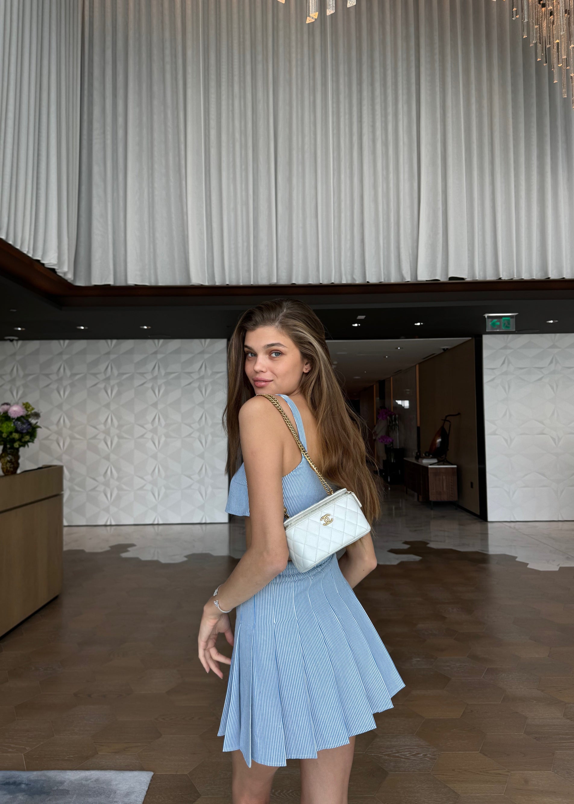 Woman in a light blue dress holding a white handbag in an indoor setting.