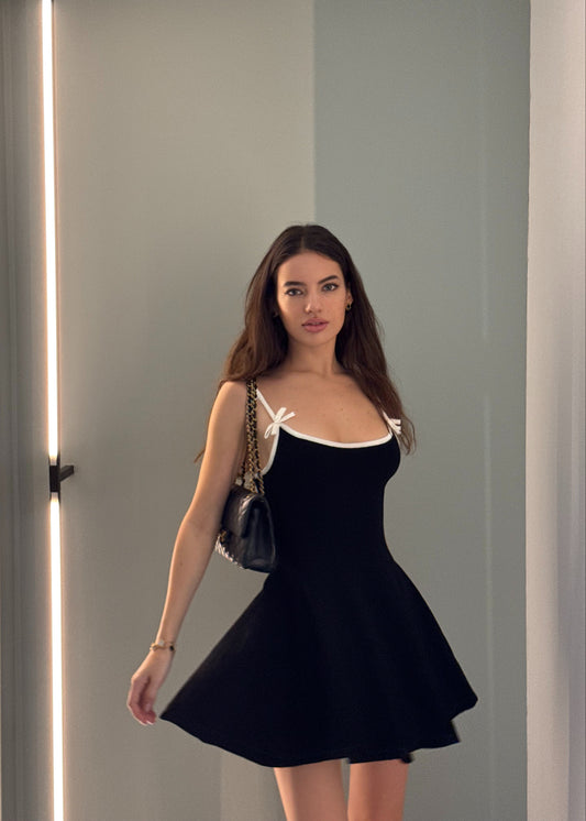 Woman wearing a black dress with a white bow detail, standing against a plain background.