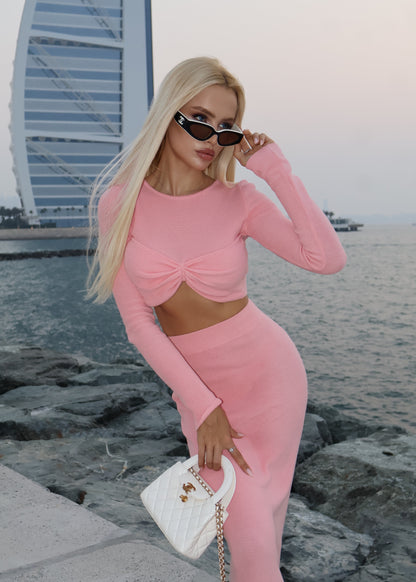 Woman in a pink outfit standing on a rocky shoreline with water and a building in the background.