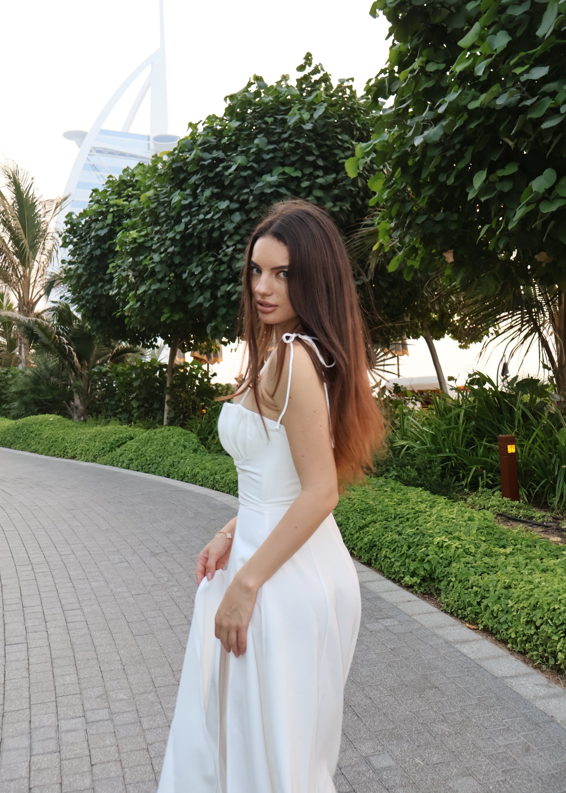 Woman in a white dress standing on a pathway with greenery and a building in the background